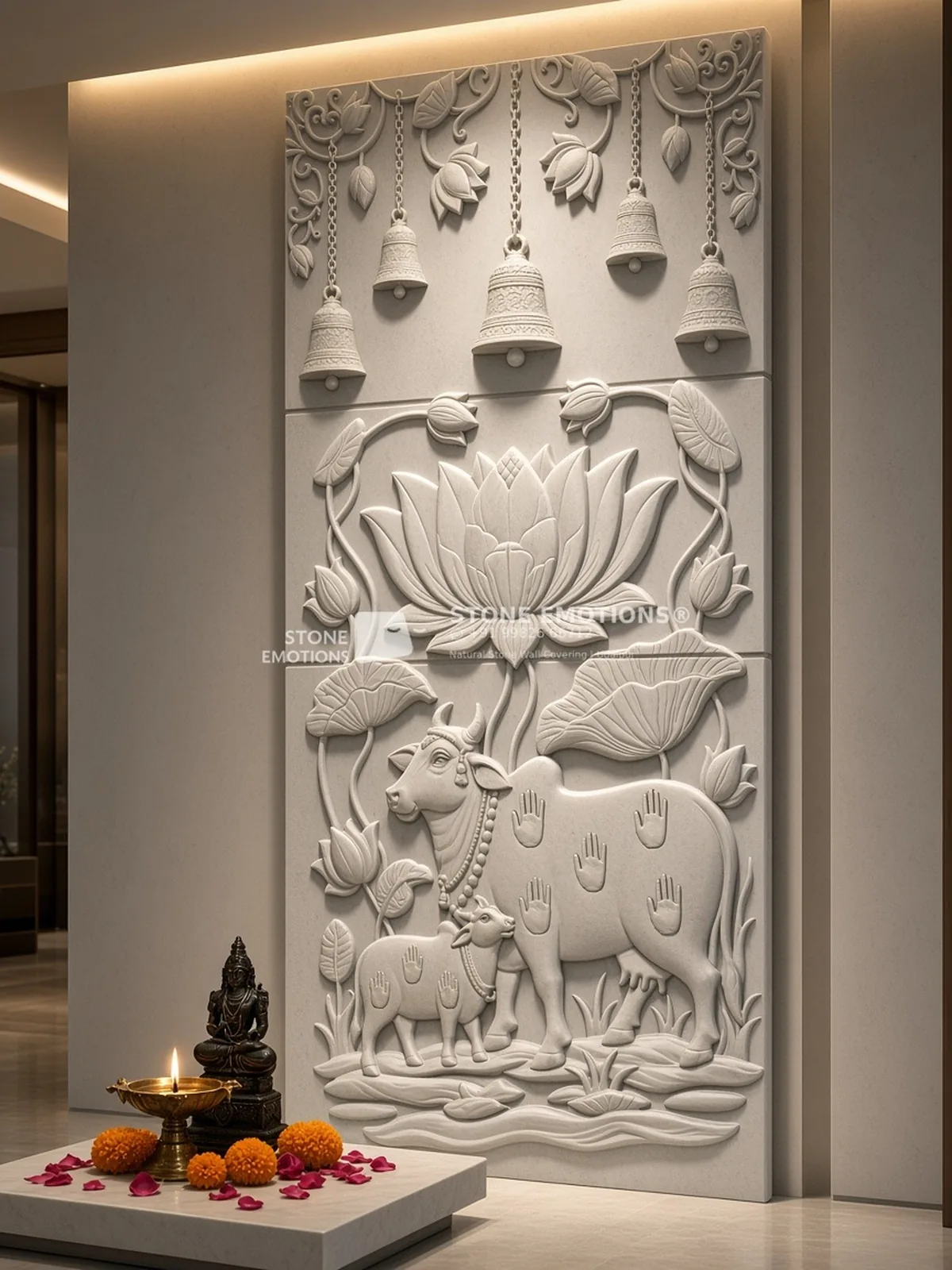 Natural stone CNC relief pooja wall panel in three stacked slabs: hanging temple bells with lotus vines, a large lotus, and Kamdhenu cow and calf on water, backlit, with brass diya and marigolds – full image